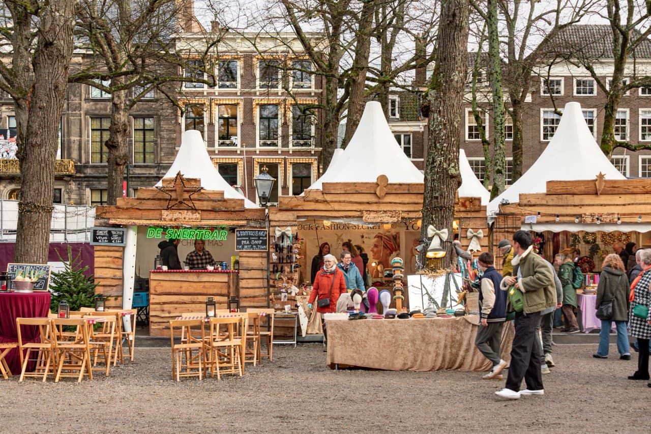 Wooden Christmas stalls with white peaked roofs under bare trees on the Lange Voorhout in The Hague, with a pea soup bar, café tables and people walking past old brick buildings.