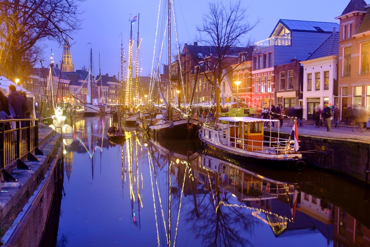 Historic sailing ships with lights strung along their masts moored in a canal at dusk, the lights reflected in the water, with people walking along the quay, brick houses and a church tower behind them.