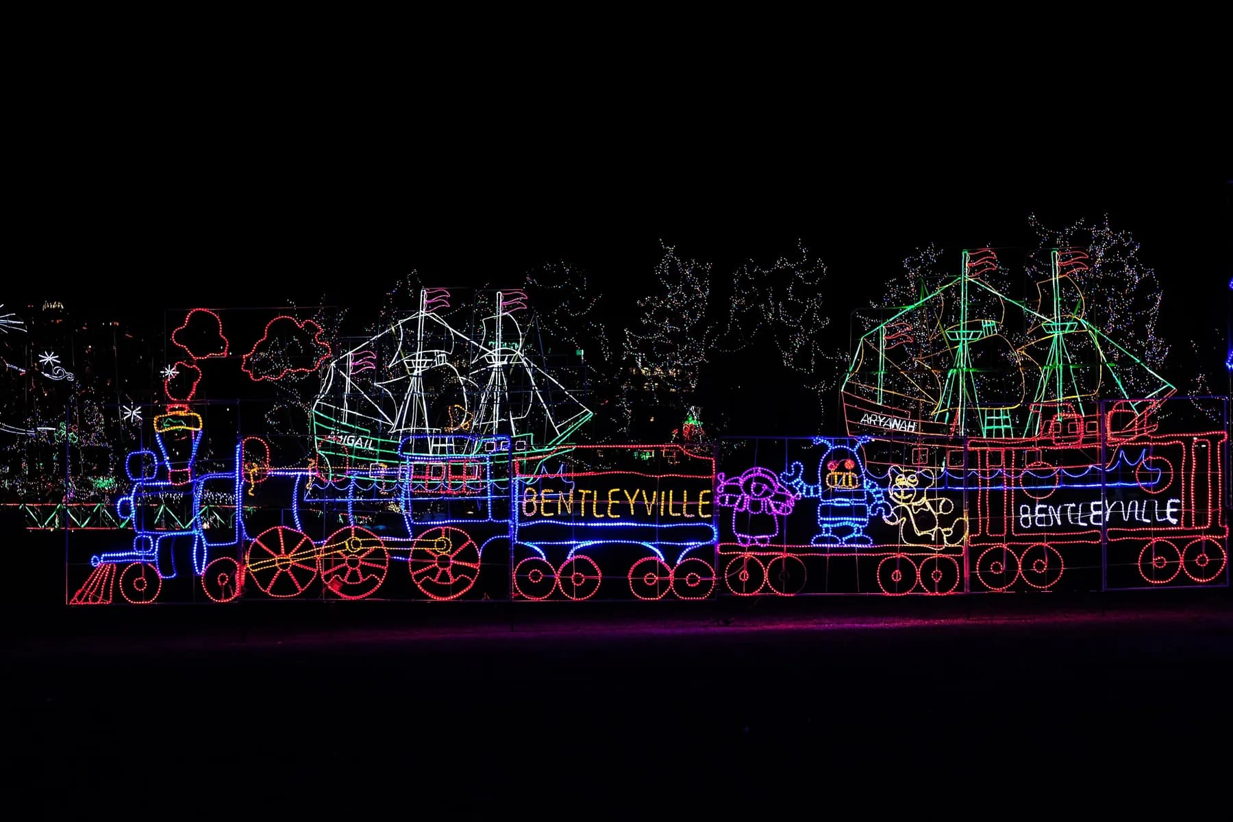 A train outlined in red, blue and yellow Christmas lights, with two lit sailing ships behind it, against a black night sky at Bentleyville in Duluth.