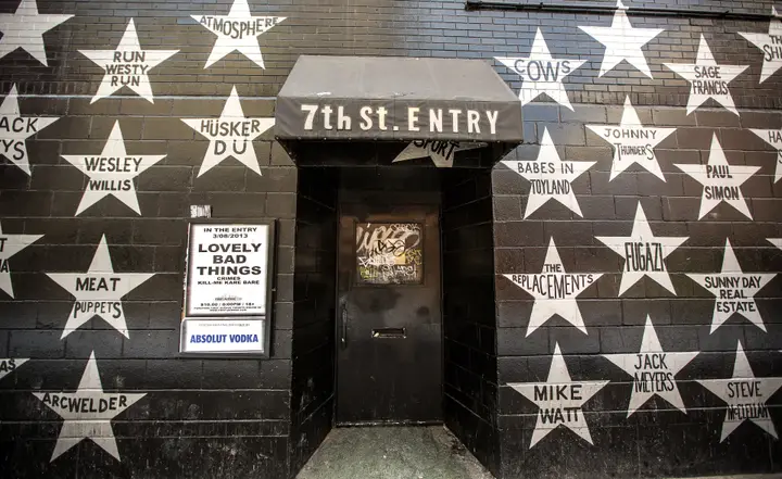 The black 7th St. Entry door and awning on 7th Street, surrounded by white stars painted with band names such as Hüsker Dü, Atmosphere and The Replacements