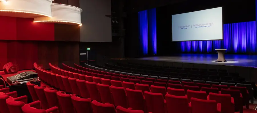 The Theatre Hall of Hanzehof seen from the side: rows of red seats and a balcony facing a stage with blue curtains and a presentation screen.