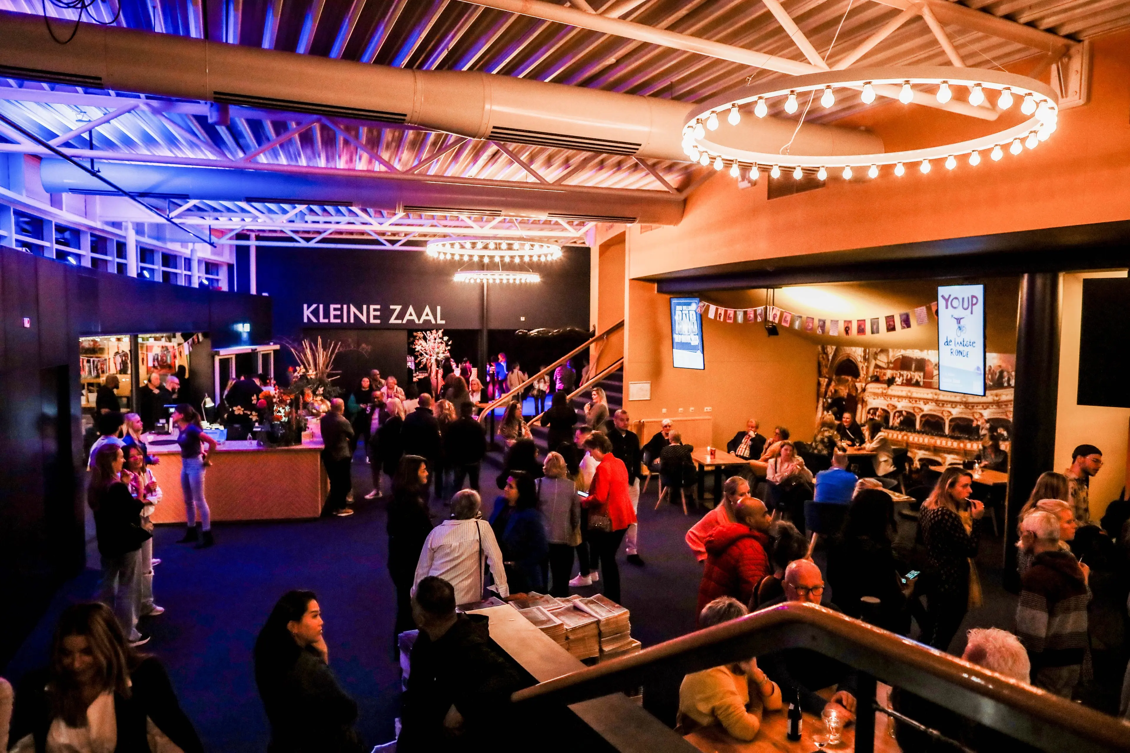 The busy foyer bar of Rijswijkse Schouwburg with the Kleine Zaal sign above the bar and visitors at tables.