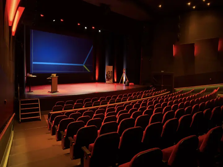 The Grote Zaal of Rijswijkse Schouwburg seen from the back, with rows of red seats facing the stage and a lectern.