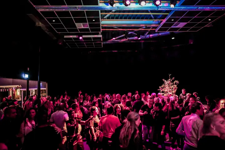 A crowd of visitors dancing in a hall of Rijswijkse Schouwburg under stage lighting, from the theatre's food and drink page.