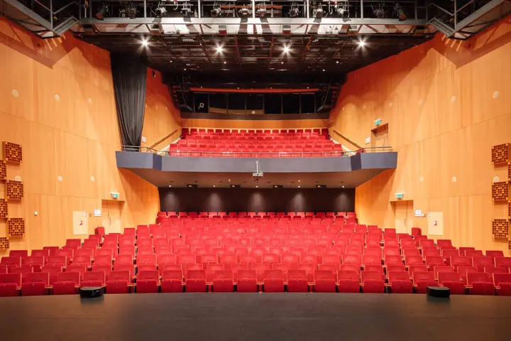 The empty theater hall of Theater de Hofnar, with red seats and a balcony, seen from the stage.