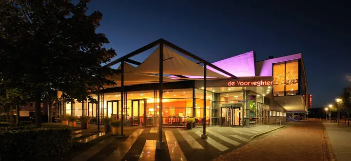 The glass front of Theater de Voorveghter at dusk, with the lit name sign above the entrance and the purple-lit façade behind.