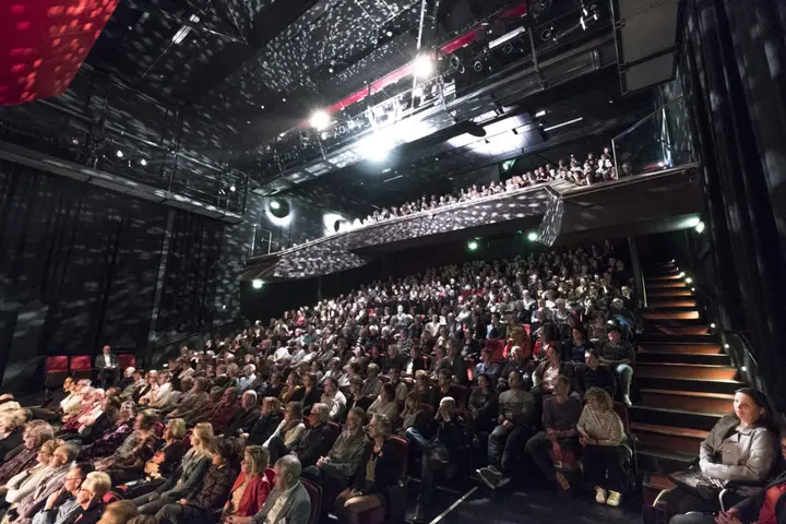 The red-seated hall of Theater de Voorveghter full of spectators during a performance, with the balcony packed above the stalls.