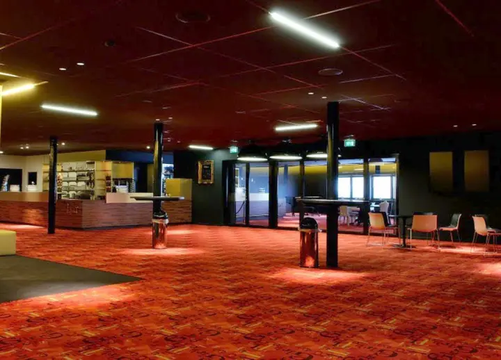 The bar and lounge of Theater Geert Teis, with a dark bar counter, high tables and a red patterned carpet.